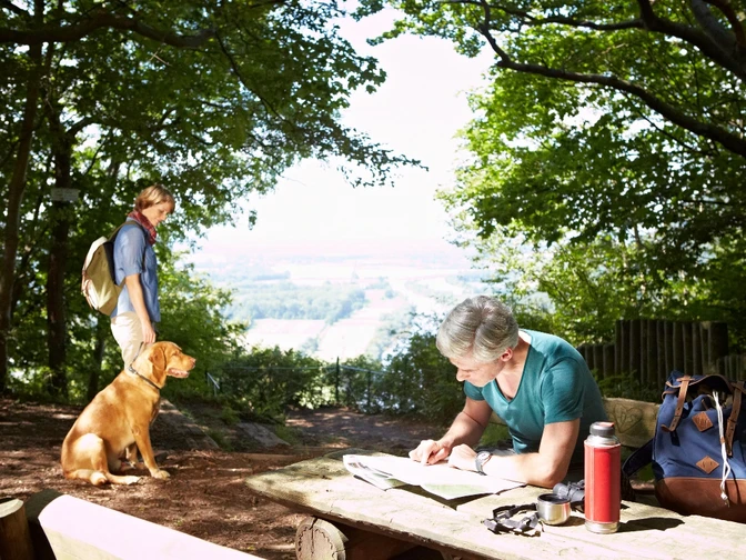 Wandern im Weserbergland Ein Wanderer rastet auf einer Lichtung im Weserbergland, blättert in einer Karte, sein Hund wartet.
