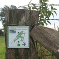 Nordic-Walking-Schild an Holzzaun zeigt Richtung in grüner Landschaft mit Stacheldrahtumzäunung.