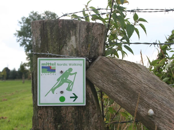 Nordic walking sign on wooden fence shows direction in green landscape with barbed wire fencing.