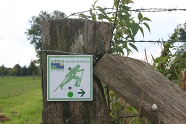NW-Schild Baumpark Nordic-Walking-Schild an Holzzaun zeigt Richtung in grüner Landschaft mit Stacheldrahtumzäunung.