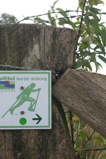 NW-Schild Baumpark Nordic-Walking-Schild an Holzzaun zeigt Richtung in grüner Landschaft mit Stacheldrahtumzäunung.