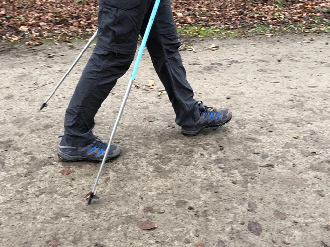 Person auf Waldweg beim Nordic Walking mit Stöcken, herbstlicher Boden, Fokus auf Beine und Schuhe.