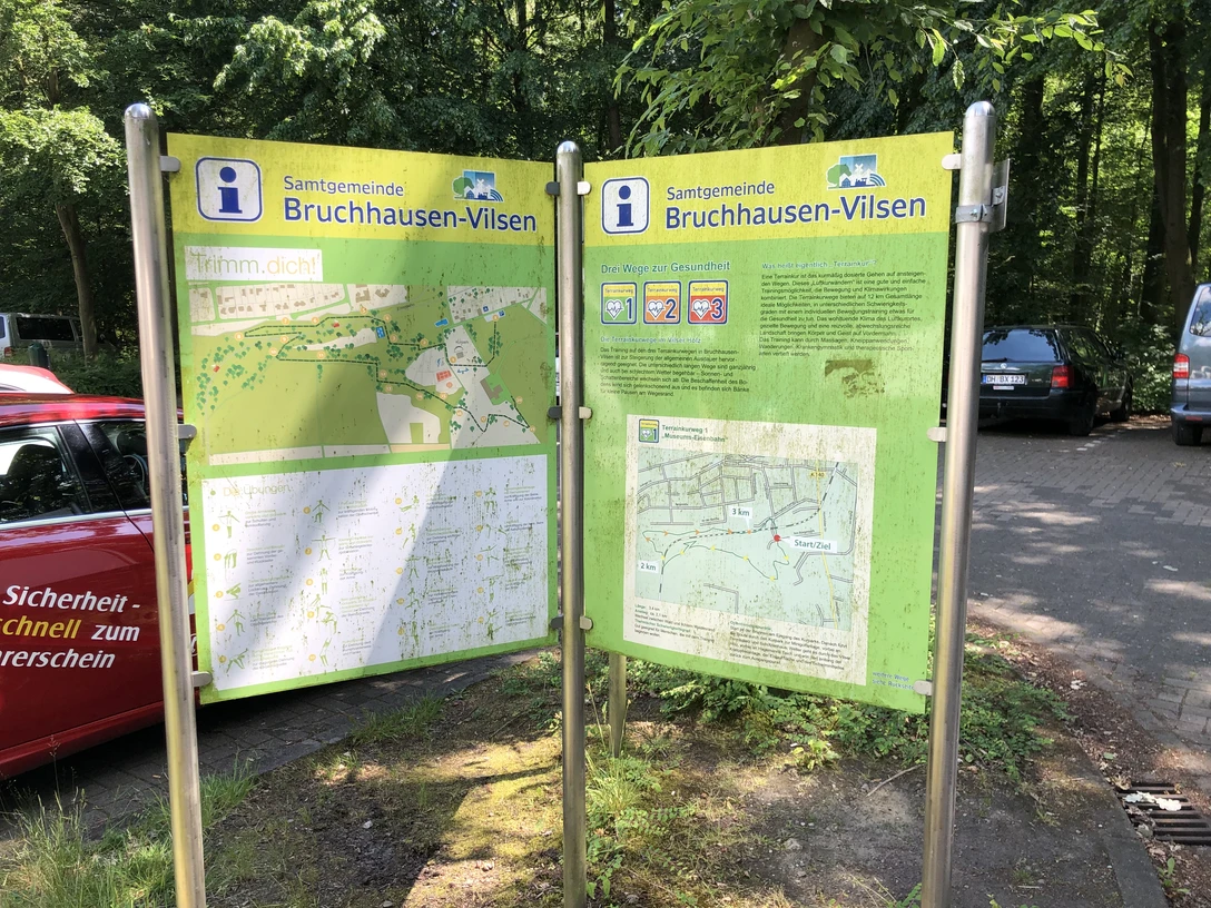 Zwei Infotafeln in Bruchhausen-Vilsen mit Wanderkarten stehen auf einem befestigten Parkplatz im Grünen.
