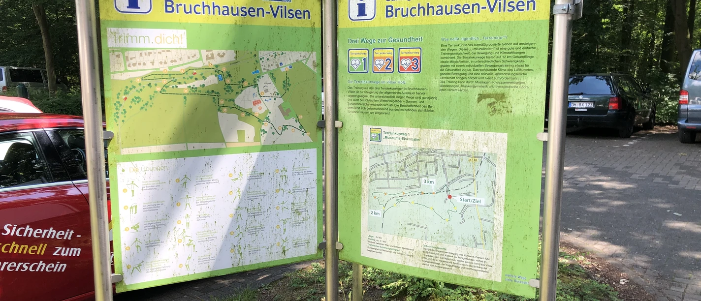 Terrainkurwege Bruchhausen-Vilsen Zwei Infotafeln in Bruchhausen-Vilsen mit Wanderkarten stehen auf einem befestigten Parkplatz im Grünen.