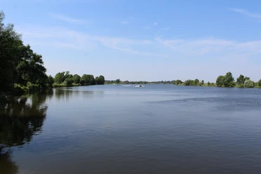 Weitläufige Flusslandschaft mit Bäumen unter blauem Himmel, ruhiges Wasser, Boote in der Ferne.