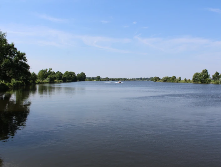 Weitläufige Flusslandschaft mit Bäumen unter blauem Himmel, ruhiges Wasser, Boote in der Ferne.