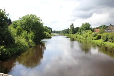 River Große Aue winds through lush countryside near Liebenau under a cloudy sky with dense vegetation.