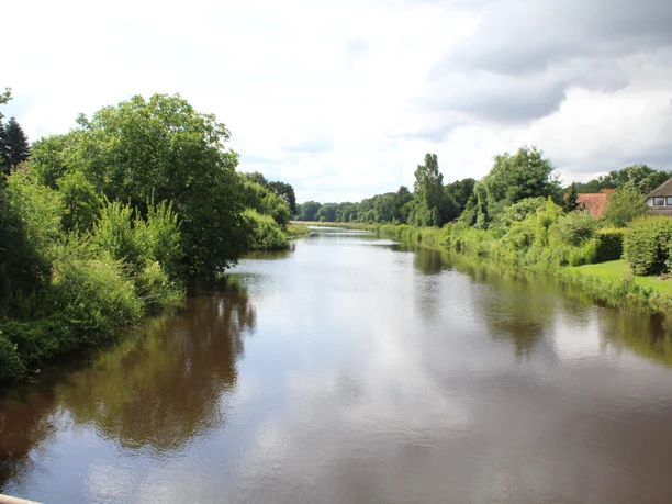 River Große Aue winds through lush countryside near Liebenau under a cloudy sky with dense vegetation.
