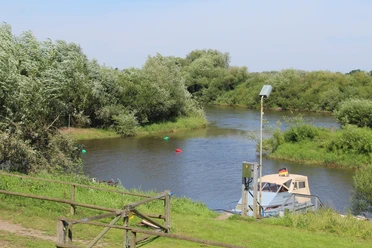 Aller bei Westen Ein Flussabschnitt mit einem Boot am Ufer, umgeben von grüner Vegetation und einem blauen Himmel.