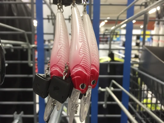 Drei kunstvolle Angelköder in rot-weiß mit Federmuster hängen an einer Metallhalterung in einem Geschäft.