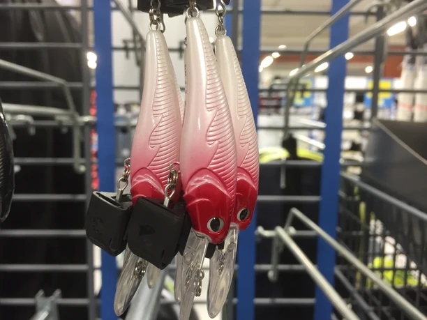 Drei kunstvolle Angelköder in rot-weiß mit Federmuster hängen an einer Metallhalterung in einem Geschäft.