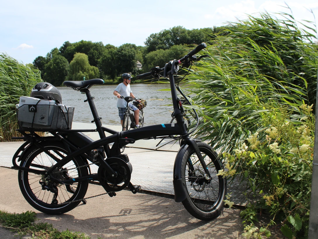 Radler Ein schwarzes Faltrad steht vor einem Flussufer, während ein Radfahrer im Hintergrund Wasser überquert.