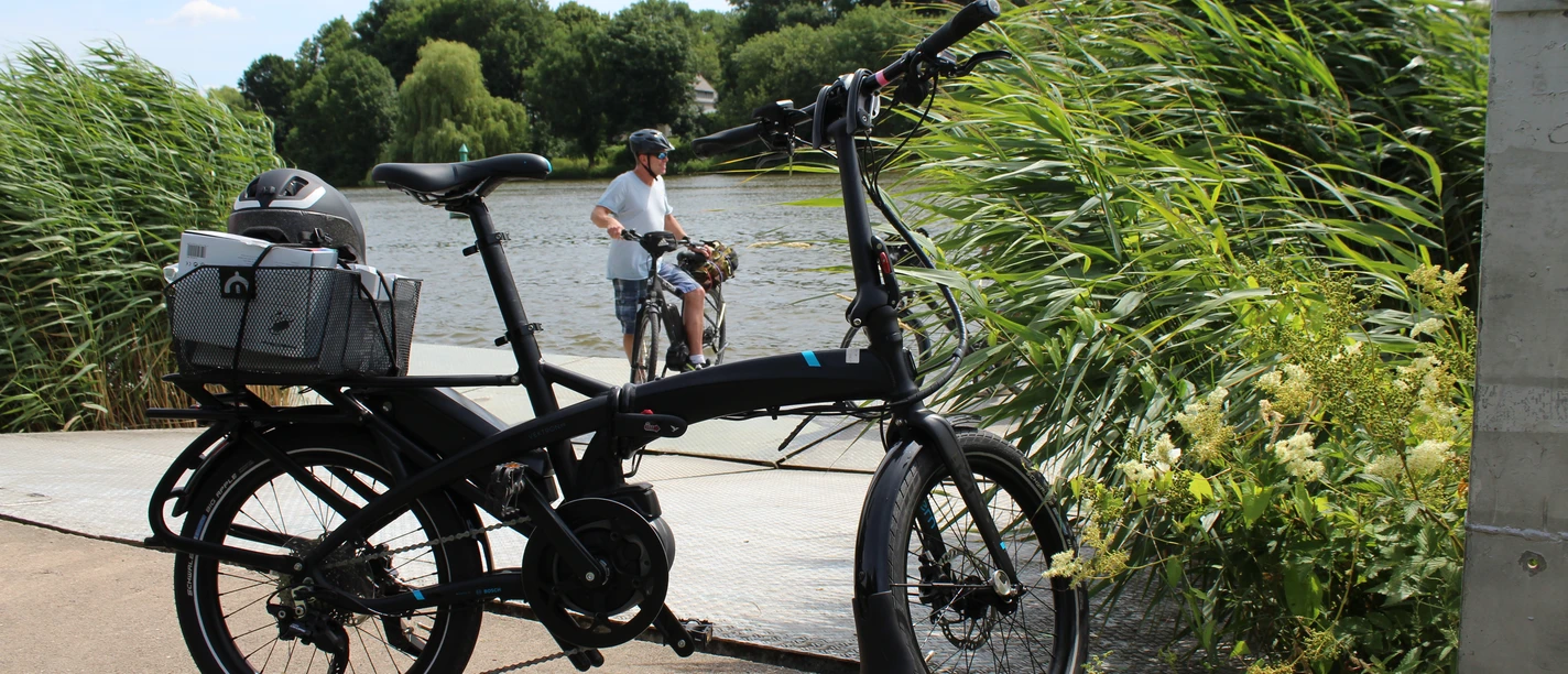 Radler Ein schwarzes Faltrad steht vor einem Flussufer, während ein Radfahrer im Hintergrund Wasser überquert.