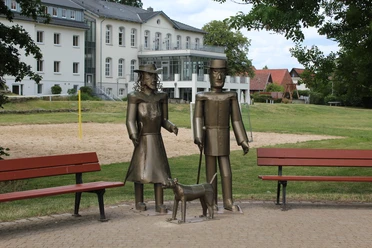 Willi, Lina und Max Drei Metallfiguren, bestehend aus zwei stehenden Personen und einem Hund, stehen auf einem gepflasterten Platz.
