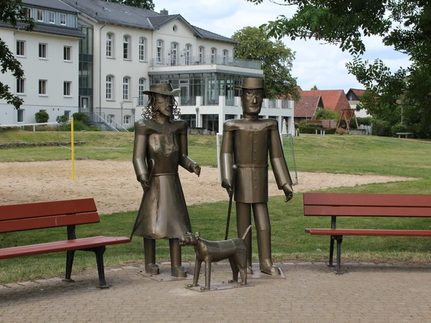 Willi, Lina und Max Drei Metallfiguren, bestehend aus zwei stehenden Personen und einem Hund, stehen auf einem gepflasterten Platz.