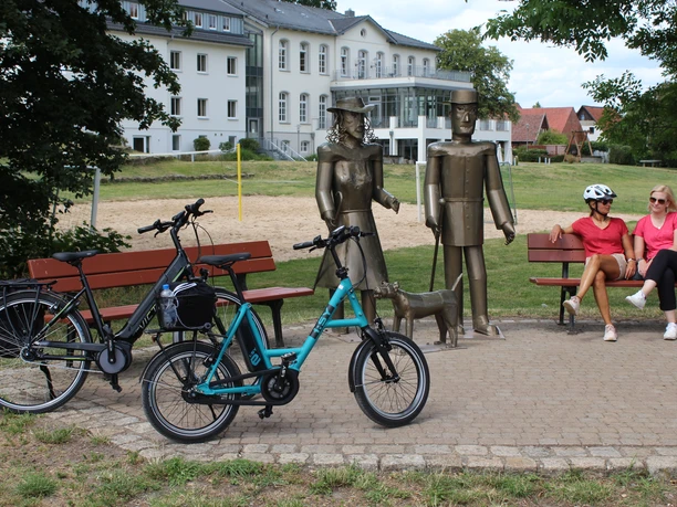 Willi, Lina und Max Skulpturengruppen aus Metall mit drei Figuren und einem Hund vor einem historischen Gebäude im Hintergrund.