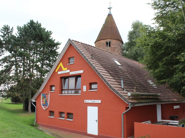 The red building with a shingle roof and a round tower in the background is surrounded by trees and a meadow.