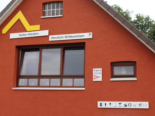 WSC-Westen Ein rotes Gebäude mit Schildern „Hafen Westen“ und „Herzlich Willkommen“, sowie einer Solatankstation.