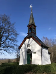 Germanakapelle <p>Weiße Kapelle mit grünem Türrahmen, markantem Turm und Wetterhahn, umgeben von Bäumen im Sonnenlicht.</p>