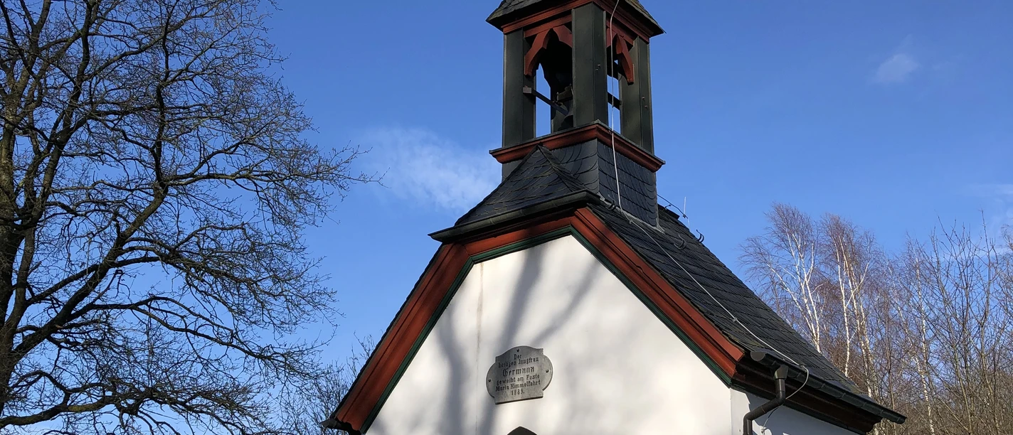 Germanakapelle <p>Weiße Kapelle mit grünem Türrahmen, markantem Turm und Wetterhahn, umgeben von Bäumen im Sonnenlicht.</p>