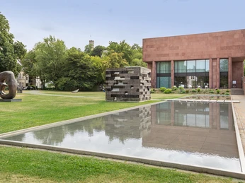 Die Skulpturen und das reflektierende Wasserbecken im Grünen vor einem modernen Museumsgebäude.