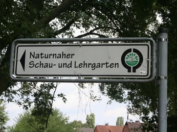 Ein Schild aus Metall weist den Weg zum naturnahen Schau- und Lehrgarten des BUND, umrahmt von Bäumen.