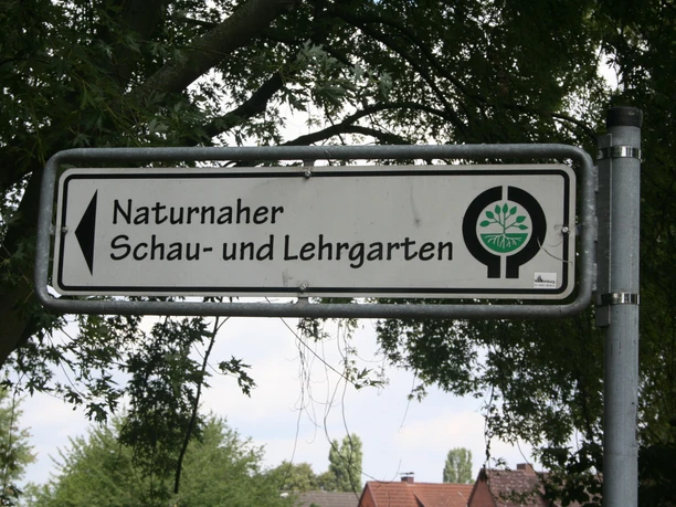 Lehrgarten BUND Ein Schild aus Metall weist den Weg zum naturnahen Schau- und Lehrgarten des BUND, umrahmt von Bäumen.