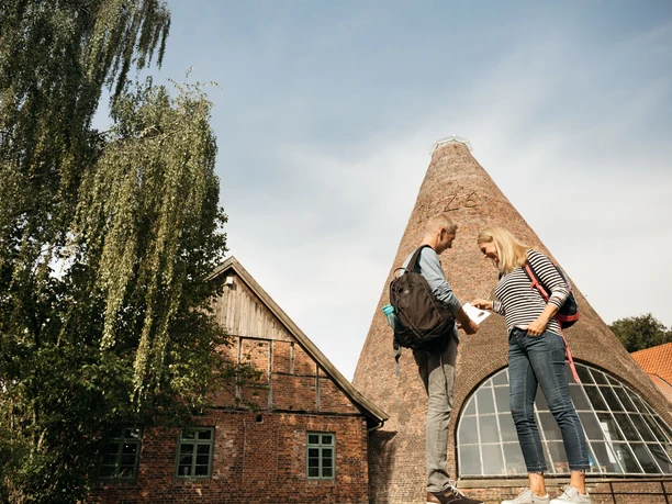 Glashütte Gernheim Historische Glashütte mit charakteristischem Konusturm, davor ein Paar mit Rucksäcken im Gespräch.
