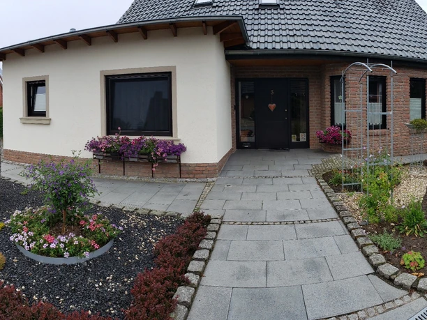 Zweistöckiges Gästehaus mit gepflegtem Garten, grauem Dach und Terrasse, umgeben von blühenden Beeten.