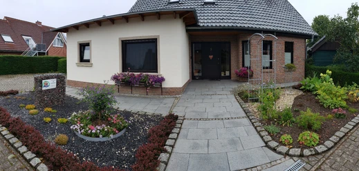 Gästehaus AM HERRMANN Zweistöckiges Gästehaus mit gepflegtem Garten, grauem Dach und Terrasse, umgeben von blühenden Beeten.