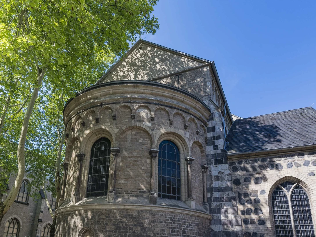 St. Cecilia Das Bild zeigt die romanische Kirche St. Cäcilien mit Rundapsis und Rundbogenfenstern unter einem klaren blauen Himmel. Die Fassade besteht aus verschiedenen Steinschichten, die Schatten der Bäume tanzen sanft darauf.The picture shows the Romanesque church of St. Cäcilien with its round apse and arched windows under a clear blue sky. The façade consists of different layers of stone, the shadows of the trees dance gently on it.