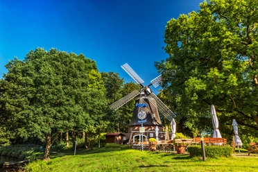 Windmühle Meppen Historische Windmühle in Meppen mit Außengastronomie, umgeben von Bäumen unter blauem Himmel.