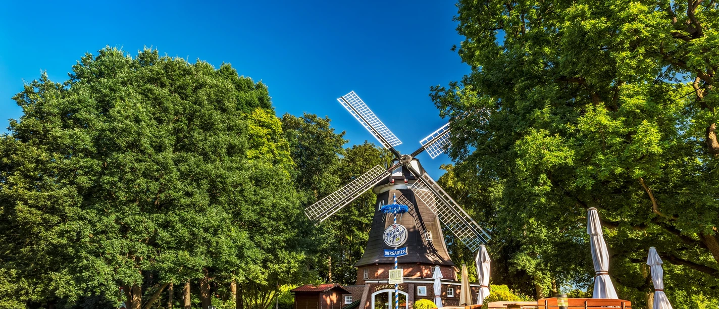 Windmühle Meppen Historische Windmühle in Meppen mit Außengastronomie, umgeben von Bäumen unter blauem Himmel.