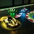Ein Mann und zwei Kinder fahren in futuristischen Karts auf einer elektrischen Rennbahn. Bunte Lichter scheinen unter den Fahrzeugen auf, während sie über eine digitale Spielfläche manövrieren. Die Strecke ist mit farbenfrohen Markierungen und Symbolen gestaltet, was das Gefühl von Videospiel-Action in Echtzeit vermittelt. Spannung und Spaß sind in der elektrisierenden Atmosphäre spürbar, während die Fahrer durch die Kurven flitzen.