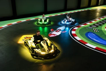 battle-kart-fahrspass.jpg Ein Mann und zwei Kinder fahren in futuristischen Karts auf einer elektrischen Rennbahn. Bunte Lichter scheinen unter den Fahrzeugen auf, während sie über eine digitale Spielfläche manövrieren. Die Strecke ist mit farbenfrohen Markierungen und Symbolen gestaltet, was das Gefühl von Videospiel-Action in Echtzeit vermittelt. Spannung und Spaß sind in der elektrisierenden Atmosphäre spürbar, während die Fahrer durch die Kurven flitzen.