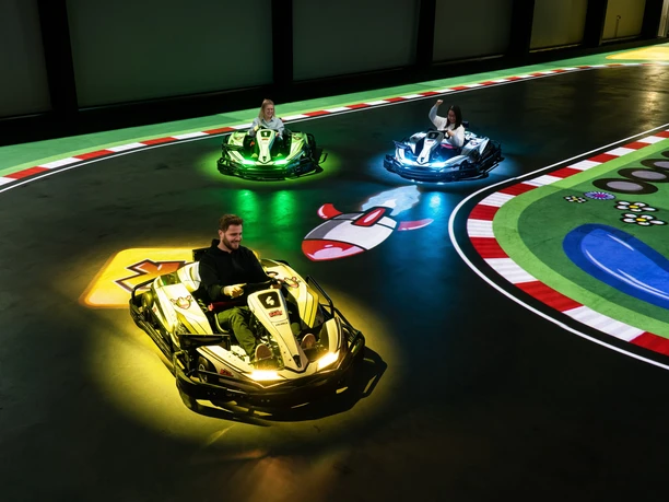 battle-kart-fahrspass.jpg Ein Mann und zwei Kinder fahren in futuristischen Karts auf einer elektrischen Rennbahn. Bunte Lichter scheinen unter den Fahrzeugen auf, während sie über eine digitale Spielfläche manövrieren. Die Strecke ist mit farbenfrohen Markierungen und Symbolen gestaltet, was das Gefühl von Videospiel-Action in Echtzeit vermittelt. Spannung und Spaß sind in der elektrisierenden Atmosphäre spürbar, während die Fahrer durch die Kurven flitzen.