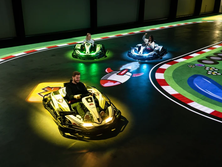battle-kart-fahrspass.jpg Ein Mann und zwei Kinder fahren in futuristischen Karts auf einer elektrischen Rennbahn. Bunte Lichter scheinen unter den Fahrzeugen auf, während sie über eine digitale Spielfläche manövrieren. Die Strecke ist mit farbenfrohen Markierungen und Symbolen gestaltet, was das Gefühl von Videospiel-Action in Echtzeit vermittelt. Spannung und Spaß sind in der elektrisierenden Atmosphäre spürbar, während die Fahrer durch die Kurven flitzen.