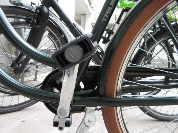 Ein schwarzes Fahrrad steht auf einem Betonboden; ein Pedal und die Kettenlöcher sind im Fokus.