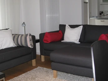Ferienwohnung Hofen: Wohnzimmer Zwei dunkelgraue Sofas mit roten und weißen Kissen neben einem beigen Teppich im Wohnbereich.