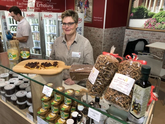 Ein Frau mit Brille steht an einem Verkaufsstand mit regionalen Produkten wie Honig und Teigwaren.
