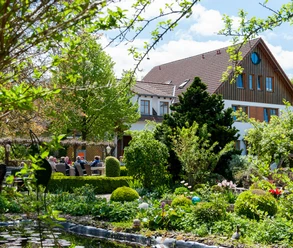 Landhaus Blumengarten: Außenansicht Üppige Gartenlandschaft mit blühenden Sträuchern umrahmt ein traditionelles Landhaus mit roten Ziegeldach.
