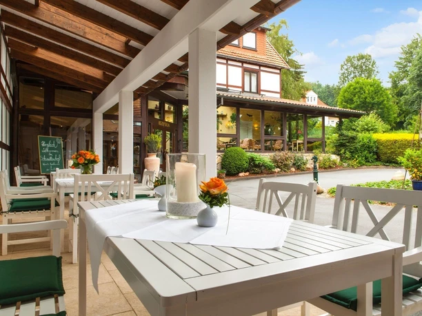 Weiß gedeckte Tische mit Kerzen und Blumen auf einer überdachten Terrasse mit Blick ins Grüne.