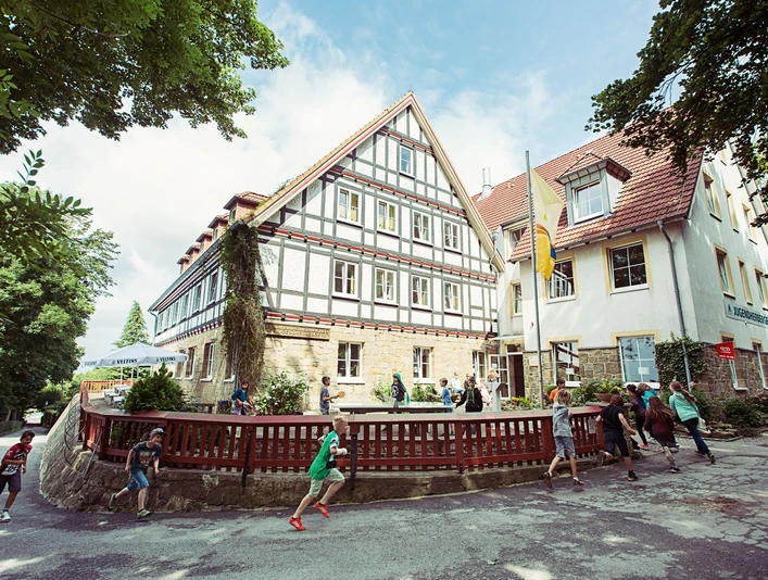 DJH Jugendherberge Tecklenburg - Außenansicht Kinder laufen vor der historischen Fassade der DJH Jugendherberge Tecklenburg. Luftige Baumkronen umrahmen die Szene.