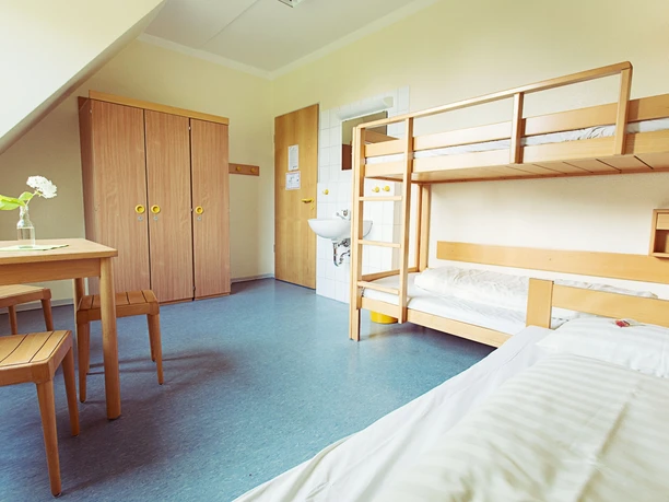 DJH Jugendherberge Tecklenburg - Zimmer Ein heller Raum mit zwei Stockbetten, kleinem Tisch, Stühlen und einem Schrank auf blauem Linoleumboden.