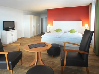 Landhotel Annelie - Doppel Komfort Ein modernes Doppelzimmer mit roter Akzentwand, Holzboden, Sitzecke und beleuchtetem Kingsize-Bett.