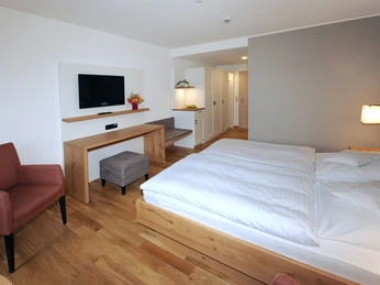 Landhotel Annelie - DZ Komfort Ein modernes Doppelzimmer mit Holzfußboden, gemütlichem Bett, Flachbild-TV und Sessel.