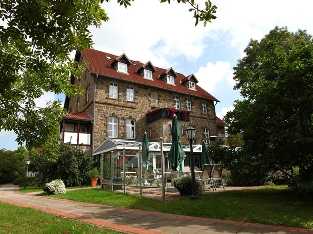 Dreistöckiges, steinernes Landhaus mit rotem Dach, Wintergarten, umgeben von gepflegten Bäumen.