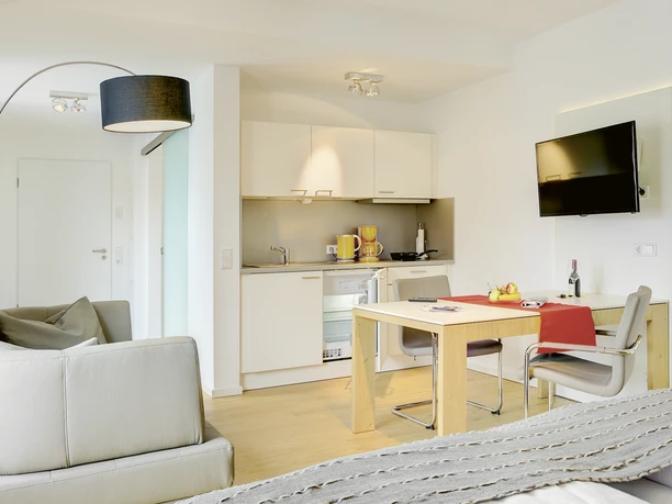 Heller, moderner Wohnraum eines Apartments mit Küchenzeile, Esstisch, Sofa und Flachbild-TV.