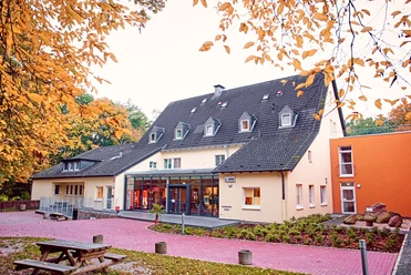 DJH Detmold - Außenansicht Jugendherberge mit mehrstöckigem Gebäude, großem Glasfoyer und Herbstlaub im Vordergrund.
