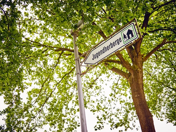 Wegweiser zu einer Jugendherberge, im Vordergrund ein Straßenschild, von Bäumen umgeben.